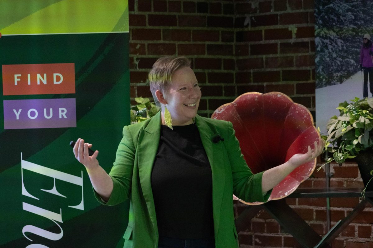 A women wearing a black shirt, green blazer and green reflective earrings stands with her hands out, palms up in the air with a smile on her face.