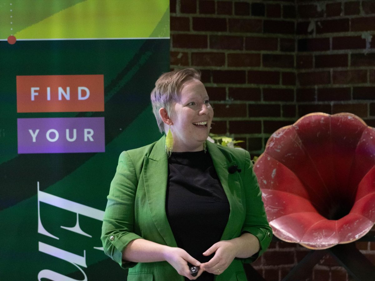 A women wearing a black shirt, green blazer, and green reflective earrings stands with her hands together, mouth slightly open with a smile.