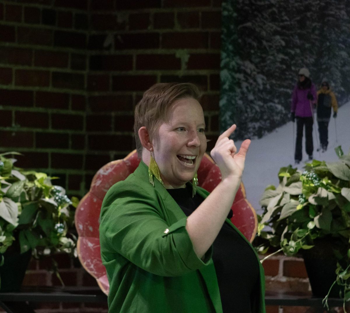 A women wearing a black shirt, green blazer and green reflective earrings with her hand up with her thumb and index finger close together. Talking with a smile on her face.