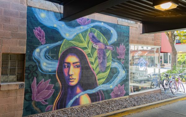 A mural on a brick wall of a woman colored with dark blue, yellow, and black. There are purple flowers on the mural as well as a bird flying behind her.