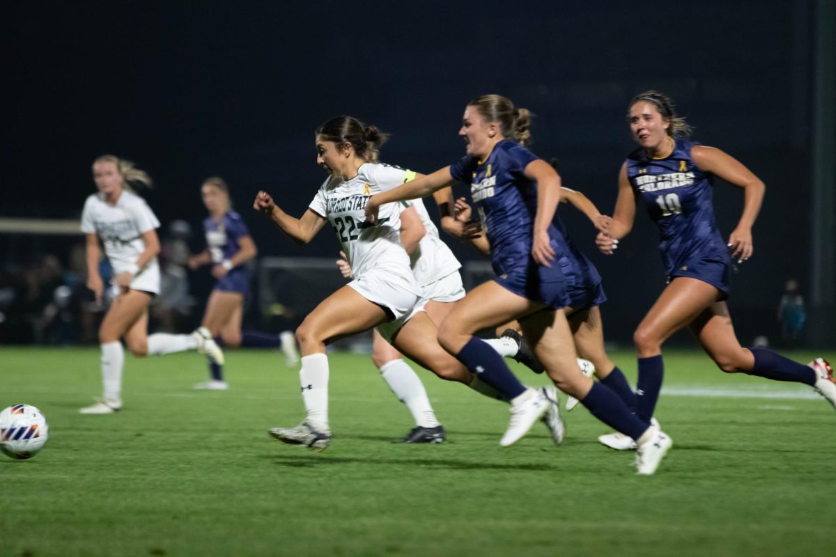Women in white and blue clothing run toward a ball in a soccer field.