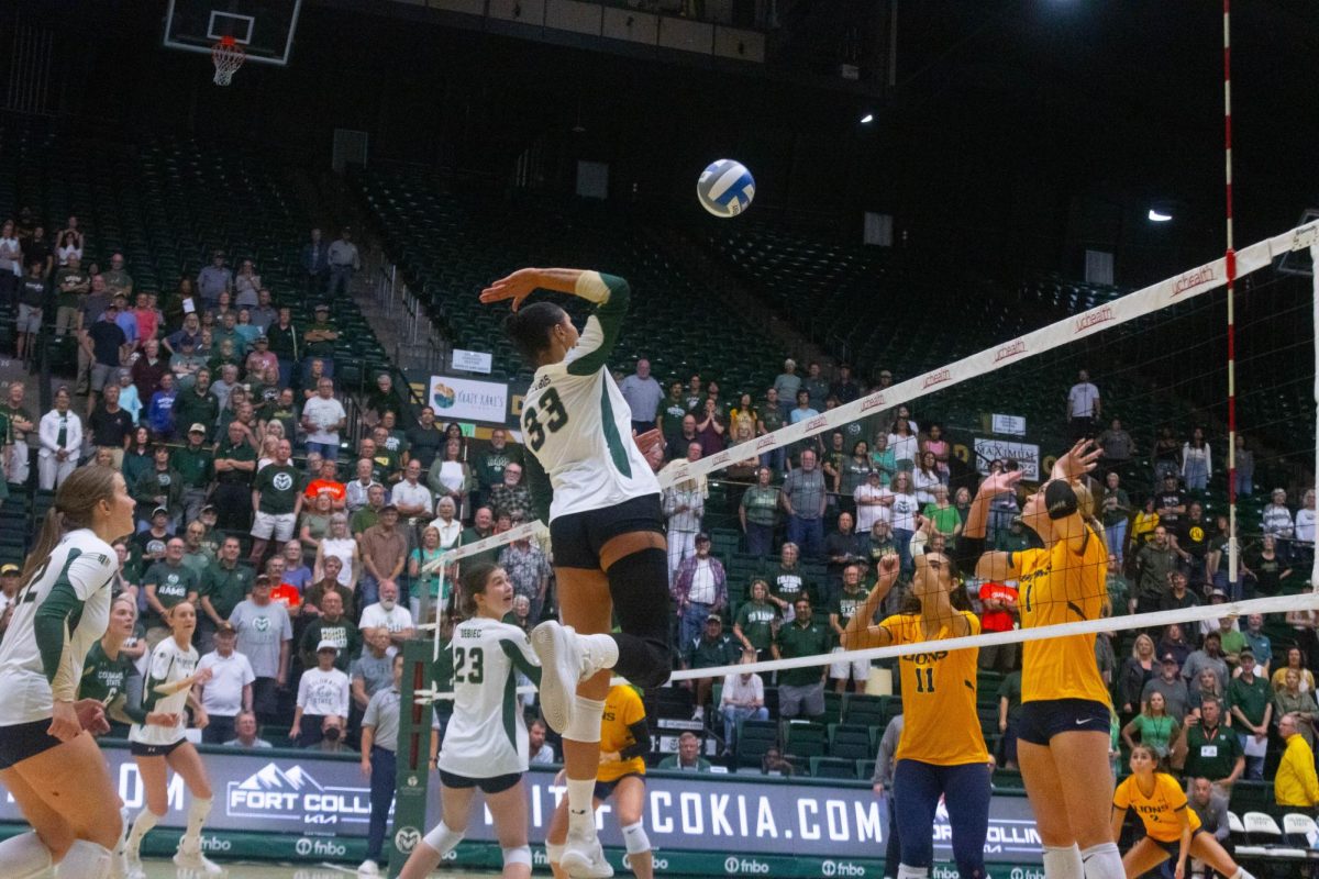 A women in a white jersey jumping above the net, preparing to spike the ball. Two women in yellow jerseys jump up as well on the other side of the net.