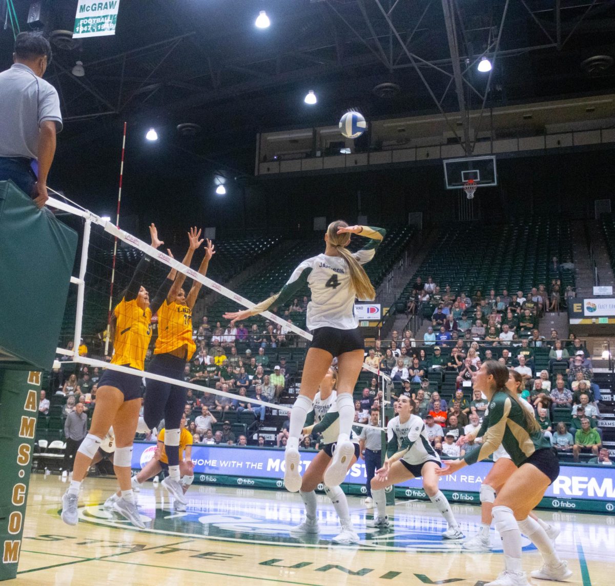 A woman in a white jersey spikes the ball, while two women in yellow jerseys are jumping to block it.