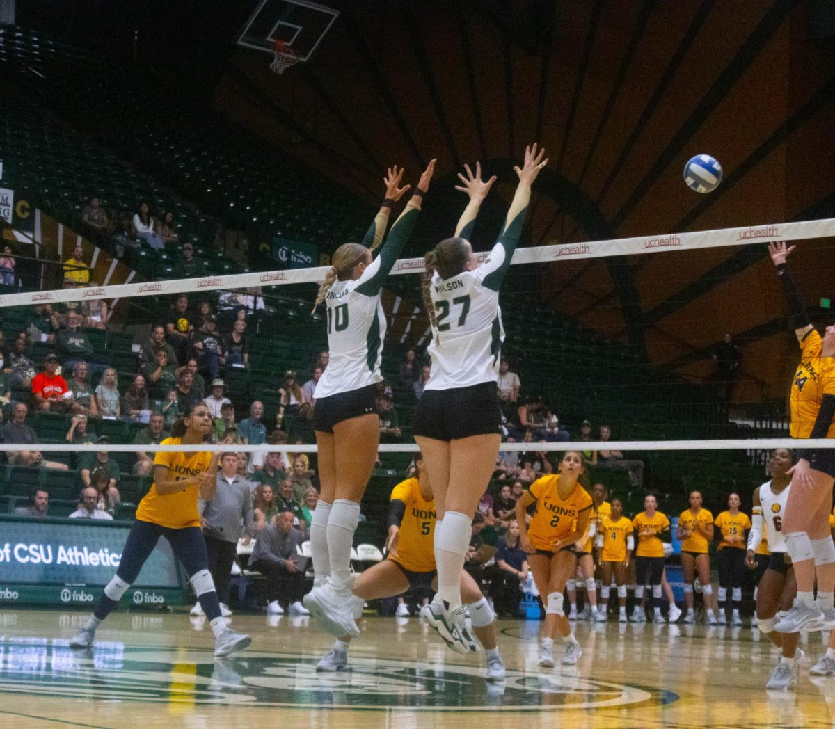 Two women in white jerseys jumping at the net. One women in a yellow jersey spikes the ball at them.