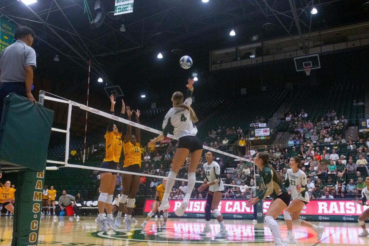 A women in a white jersey jumps high to hit the ball. Two women in yellow jerseys jump at the other side of the net.