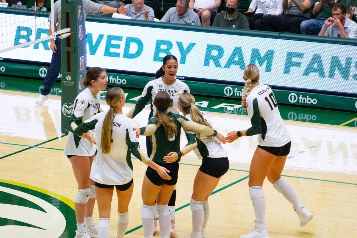 5 women in white jerseys and one woman in a green jersey huddle together. All of them are smiling.