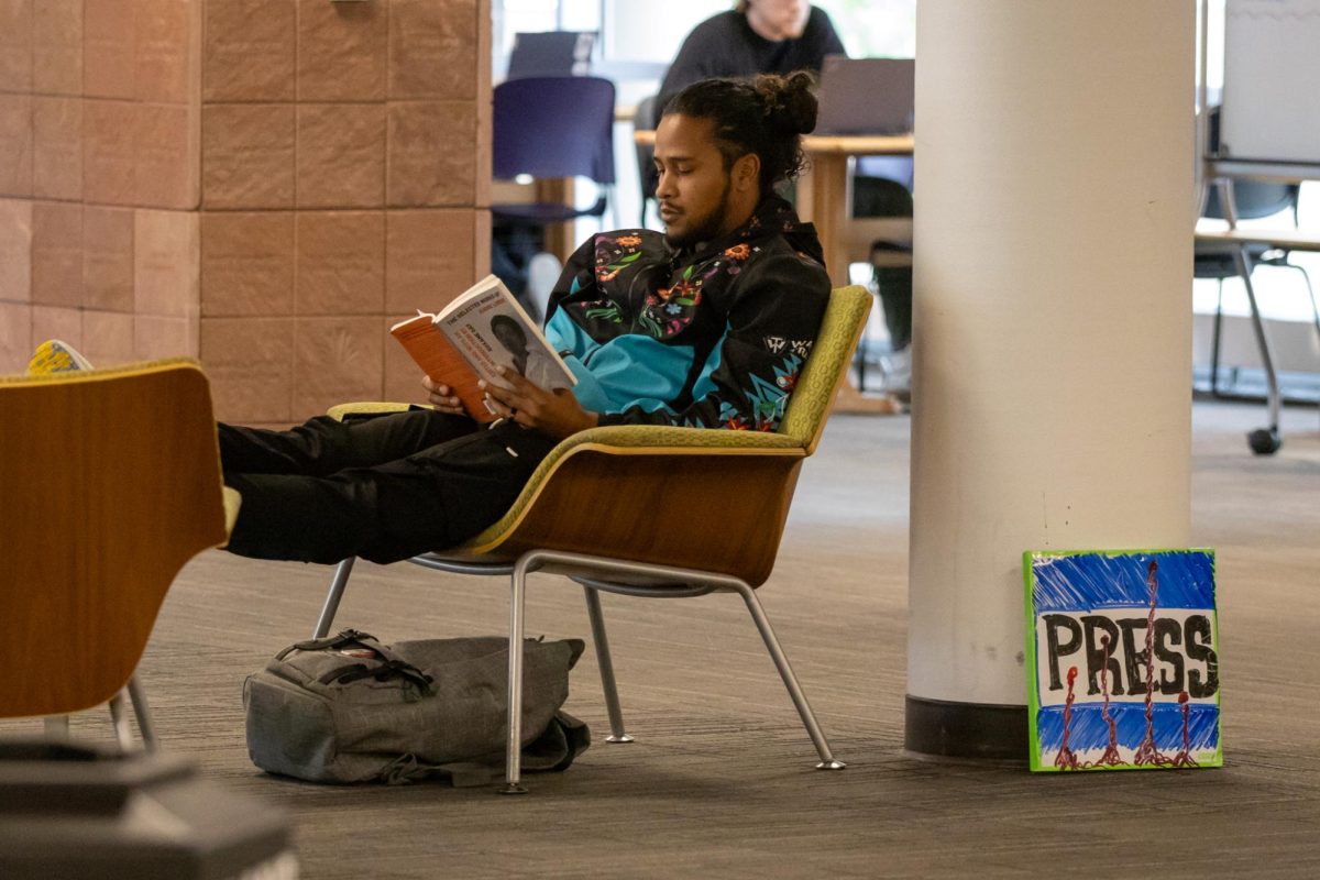 A young man sits in a chair reading while a whiteboard sign that has the word “PRESS” drawn in expo sits on the floor next to him.