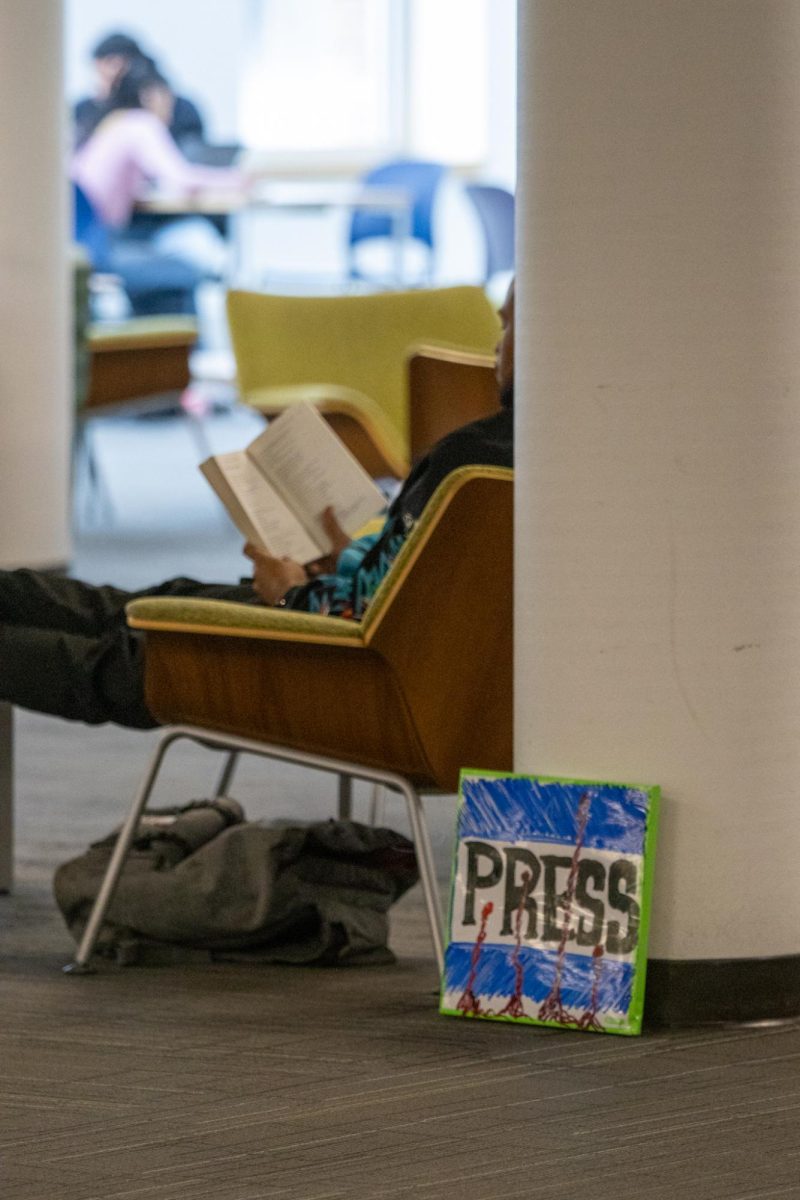 A young man sits in a chair reading while a whiteboard sign that has the word “PRESS” drawn in expo sits on the floor next to him.