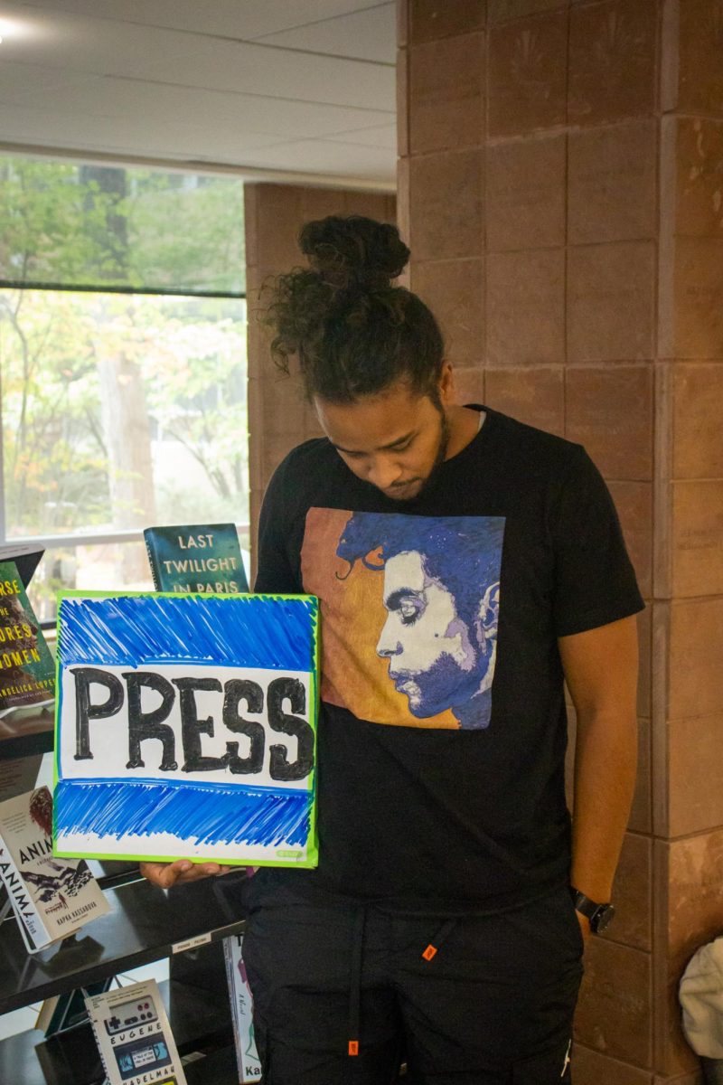 A young man posing for a picture, holding a whiteboard sign that has the word “PRESS” drawn in expo.