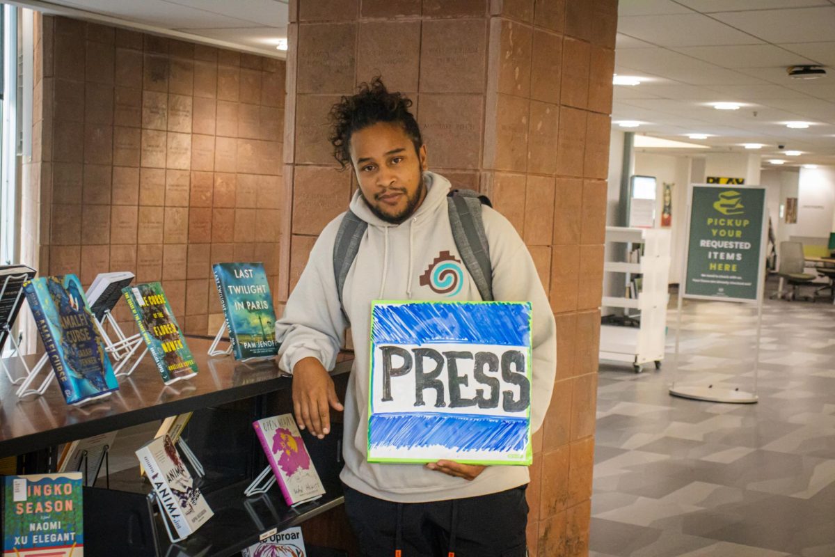 A young man posing for a picture, holding a whiteboard sign that has the word “PRESS” drawn in expo.