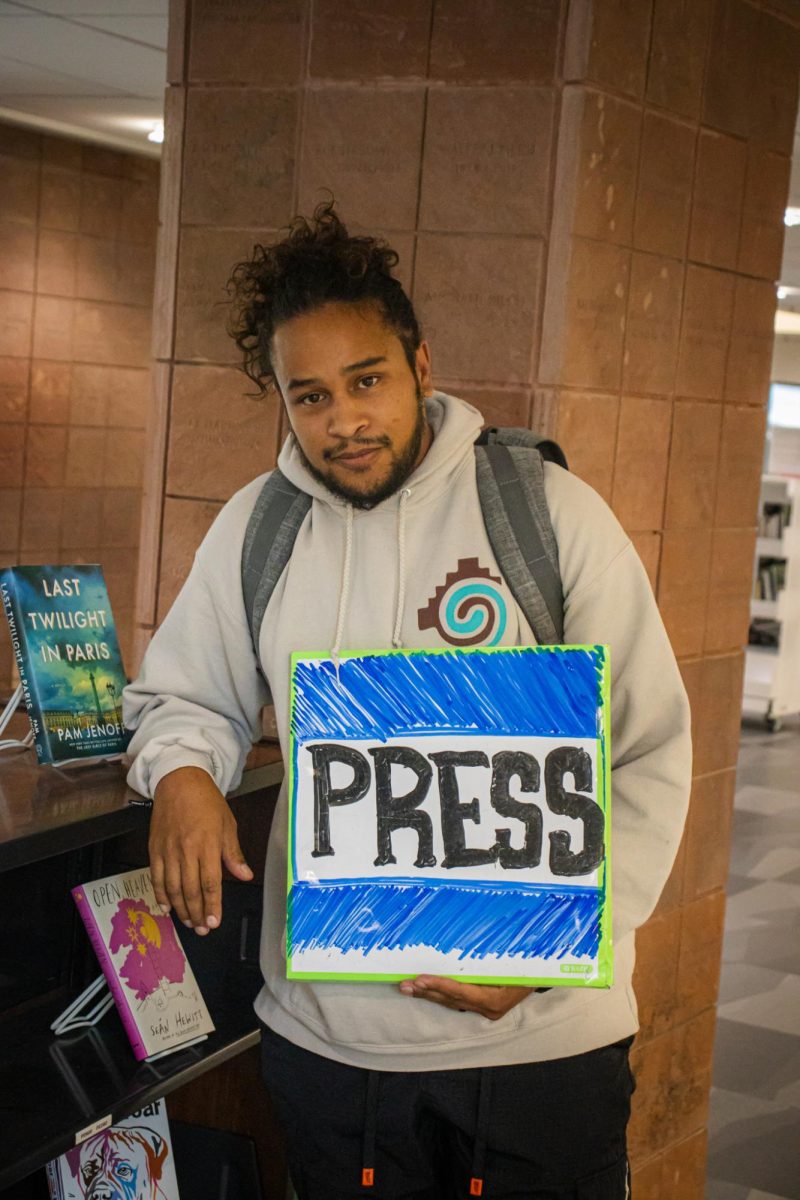 A young man posing for a picture, holding a whiteboard sign that has the word “PRESS” drawn in expo.