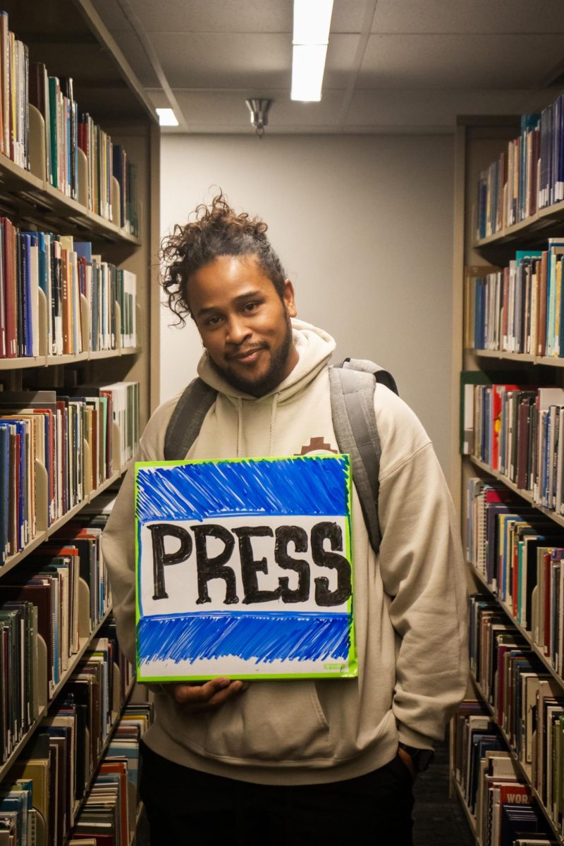 A young man posing for a picture, holding a whiteboard sign that has the word “PRESS” drawn in expo.