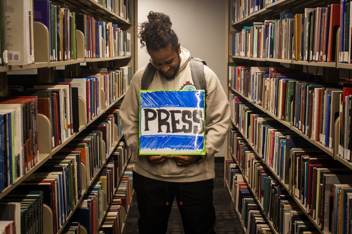 A young man posing for a picture, holding a whiteboard sign that has the word “PRESS” drawn in expo.