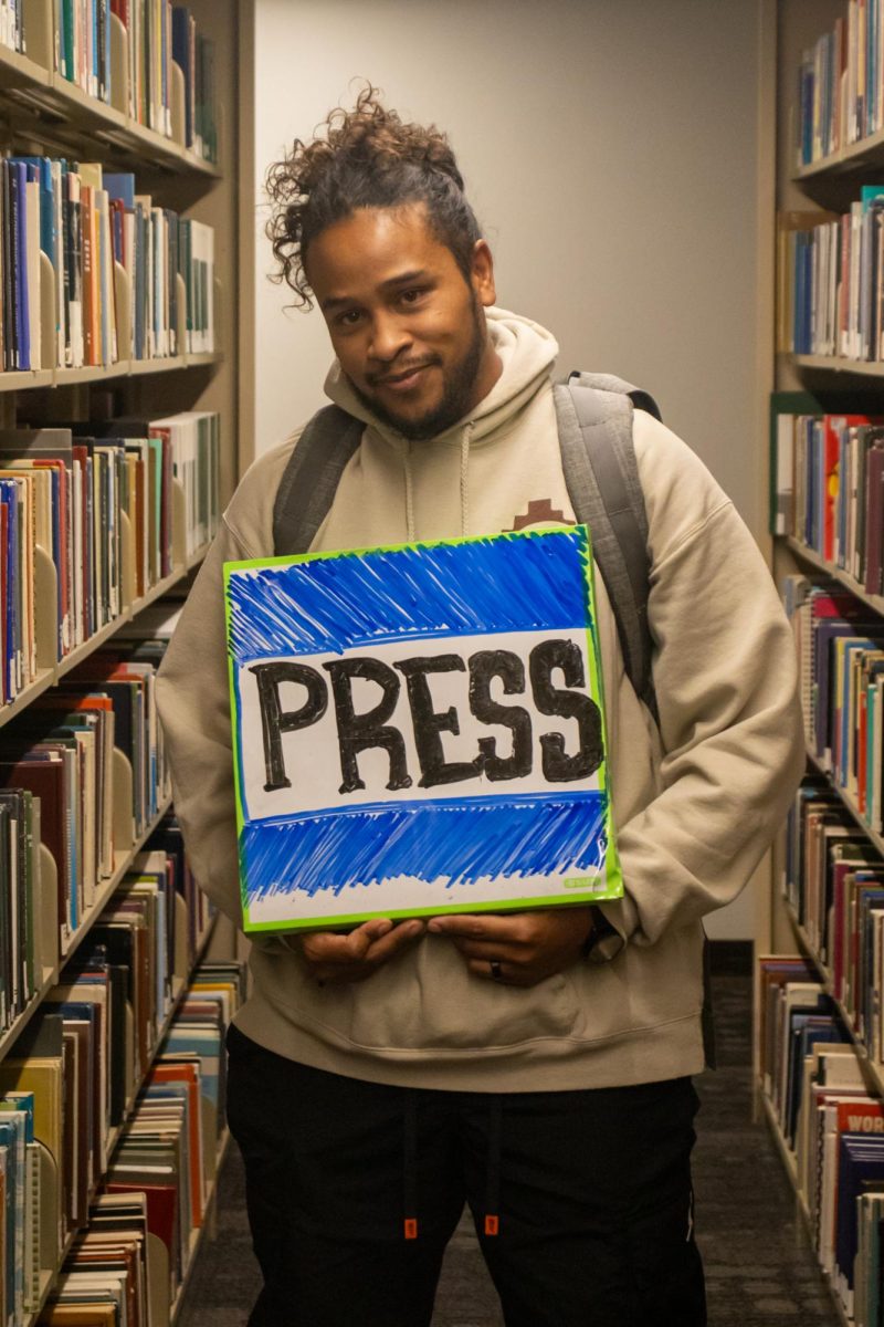 A young man posing for a picture, holding a whiteboard sign that has the word “PRESS” drawn in expo.