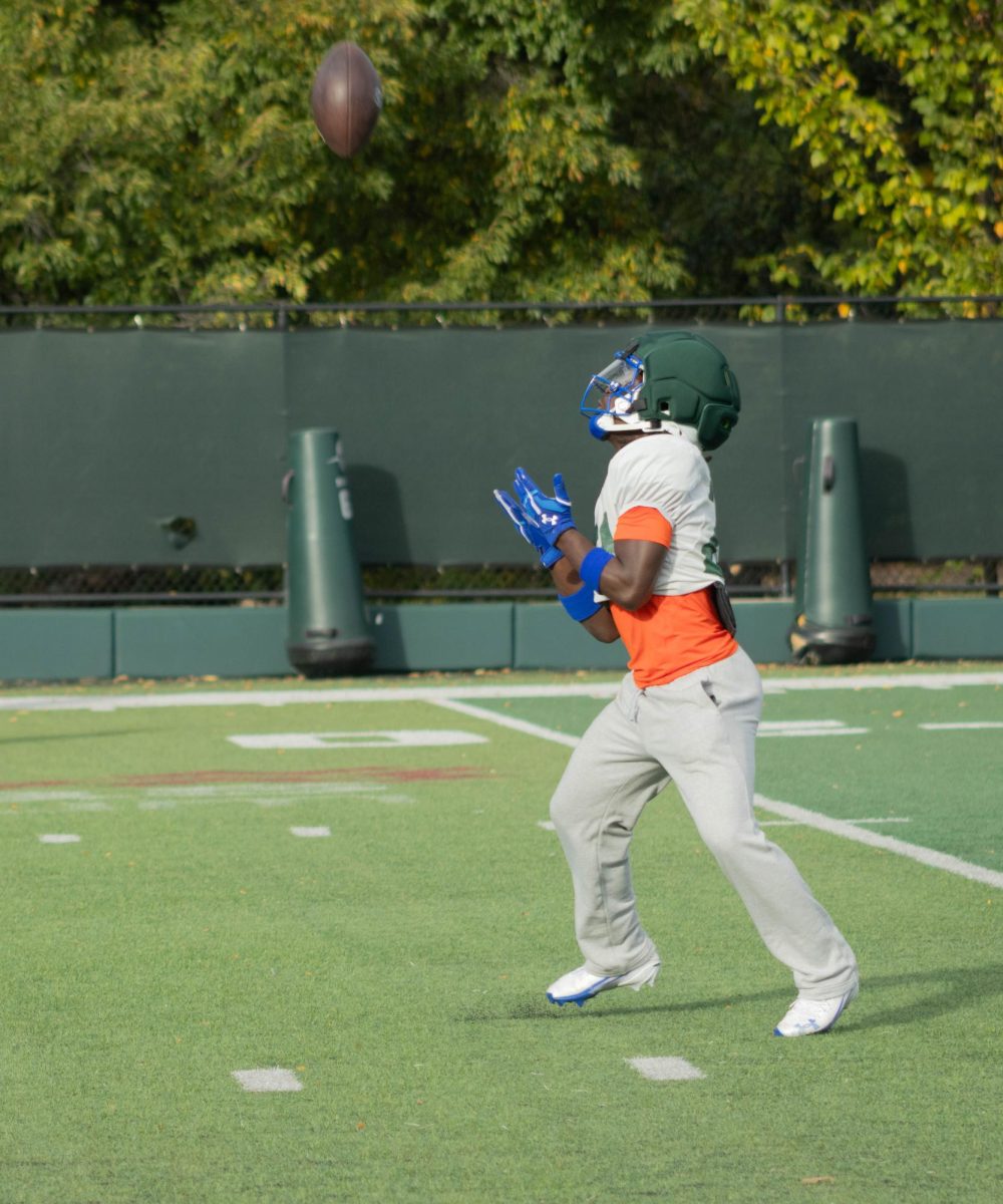 A player wearing an orange undershirt and grey sweatpants looks up and holds his hands out below the football.