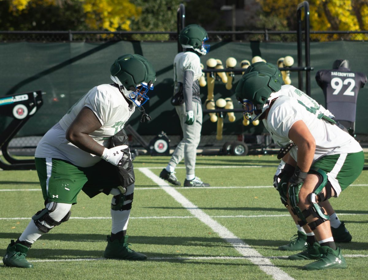 Three players with their knees slightly bent over next to each other. There is one player on the left and two on the right. The player on the left is holding a padded bag in front of him.