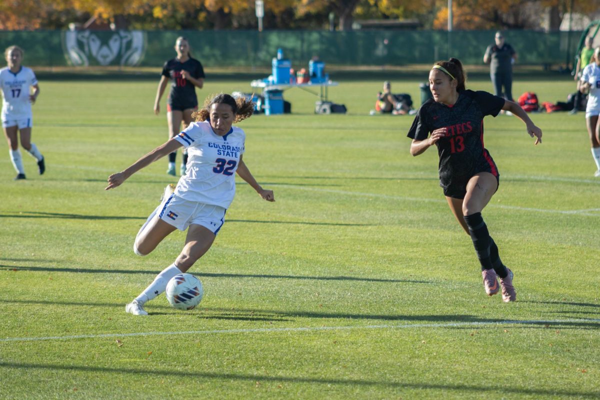 A women wearing a white jersey extends her right leg at the ball in front of her. There is a women in a black jersey on her right running at her.
