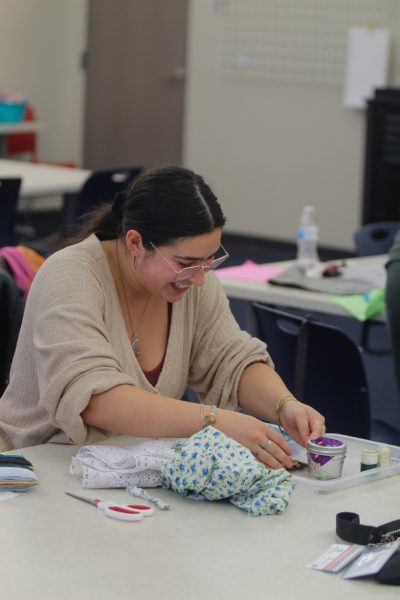 A smiling woman with dark hair, glasses, and a cream cardigan places brown buttons in a plastic dish. On the table is a pair of scissors, and patterned fabric.