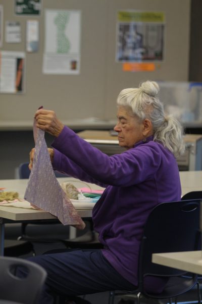 An older woman with grey hair is wearing a purple fleece and holding purple patterned fabric out in front of her.