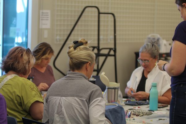 A group of middle aged women sit around a table while looking at their individual projects. The woman closes to the camera holds an embroidery hoop with light blue fabric.