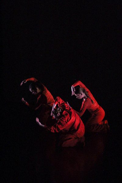 Three dancers in red strike a pose on the ground during their performance. They sit on their knees and face to the left while holding an arm above their head.