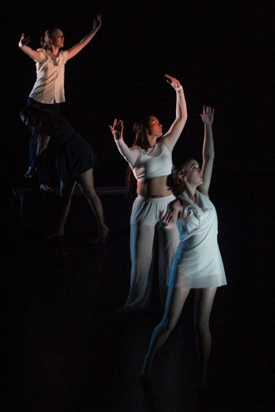 Three dancers in different white outfits stand with their arms over their head and look gently to the right. Another dancer in darker clothes stands lower and faces to the left.