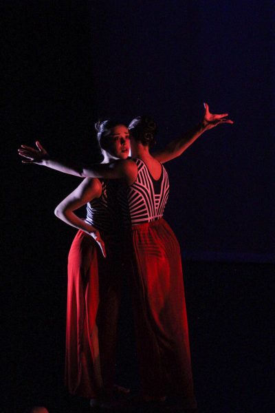 Two dancers in striped tops and red skirts embrace each other while extending their right arm. One dancer faces the camera, and another faces away.