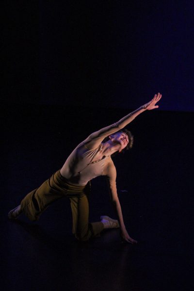 A male dancer strikes a pose on the ground while reaching upwards. His face is turned towards his hand.