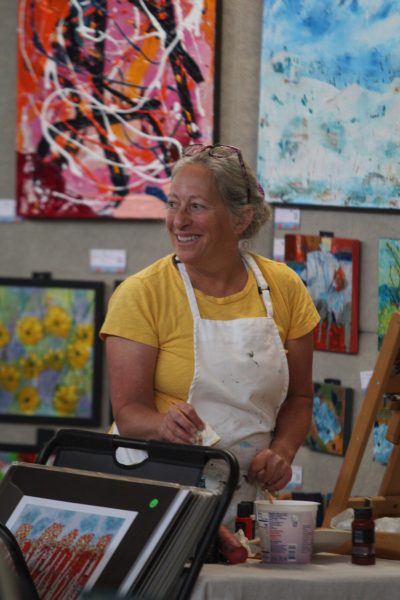 A woman with grey hair, a yellow shirt, and a white apron stands in behind an easel and smiles to a person standing out of frame. Behind, her artwork is visible.