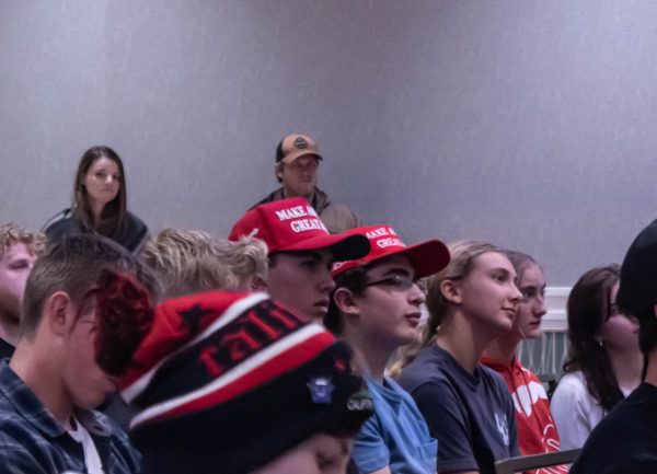 A crowd of people sitting down in chairs look up at the podium. Two men are wearing red hats that say "Make America Great Again"