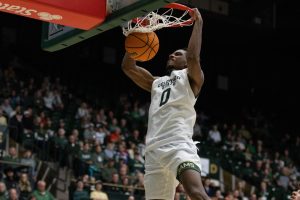 A man in an all white jersey hangs from the rim with both arms flexed, he has an angry expression on his face. The ball is falling through the rim.
