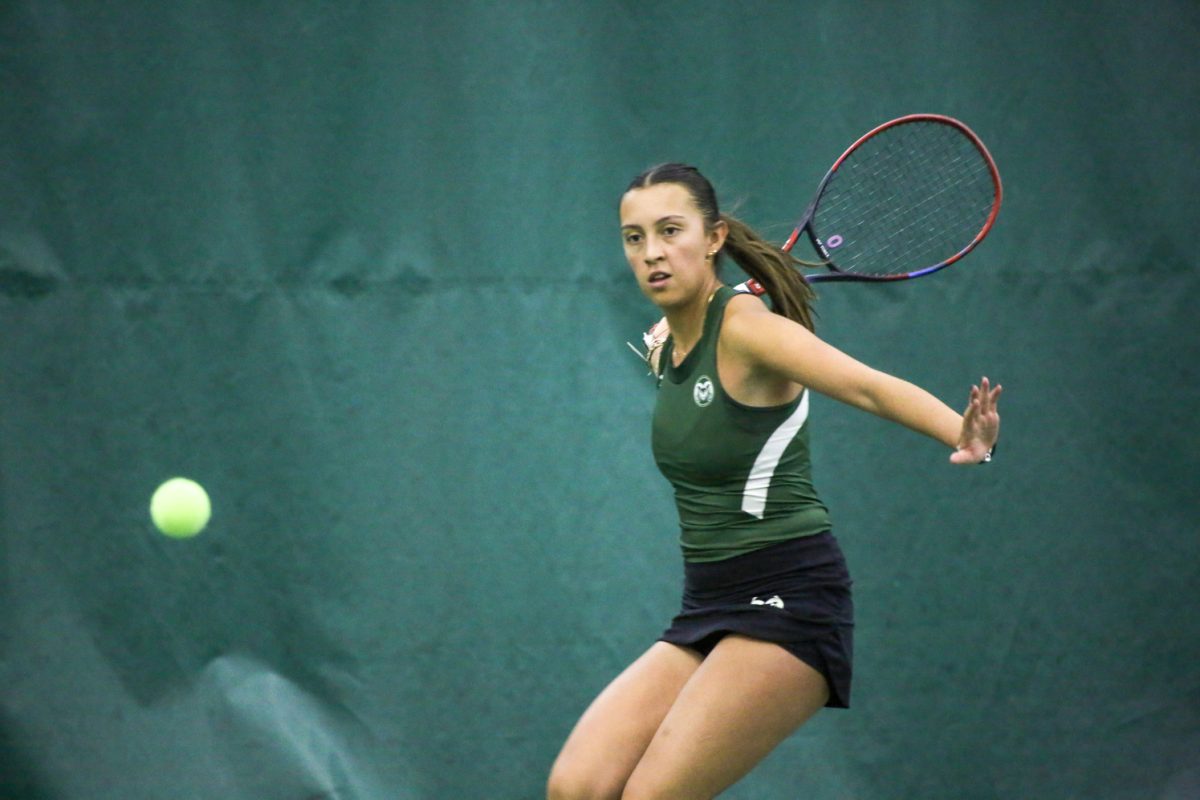 A woman in a green shirt and black skirt holds a racket above her head in front of a green wall.