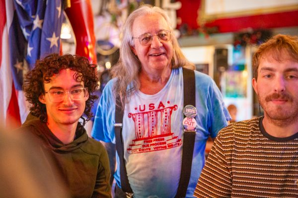 A man with long hair wears suspenders and buttons are attached stands between two men.