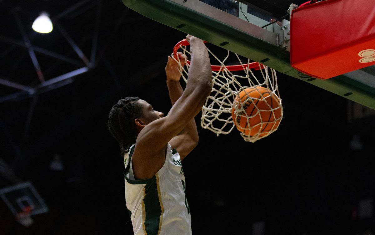 A man wearing an all white jersey has both of his hands on the rim as the ball is caught in the net moving downward.