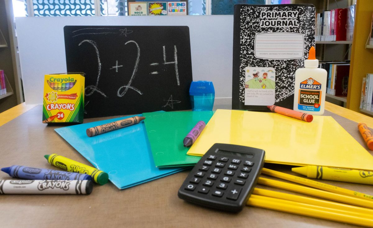 School supplies laid out in an appealing manor, there is a black chalkboard with "2+2=4" written on it, a black composition book to the right of the chalkboard, a box of crayons is stood up leaning on the chalkboard, and a bottle of glue is leaning on the composition book. In front of everything is three colored folders with crayons littering the photo.