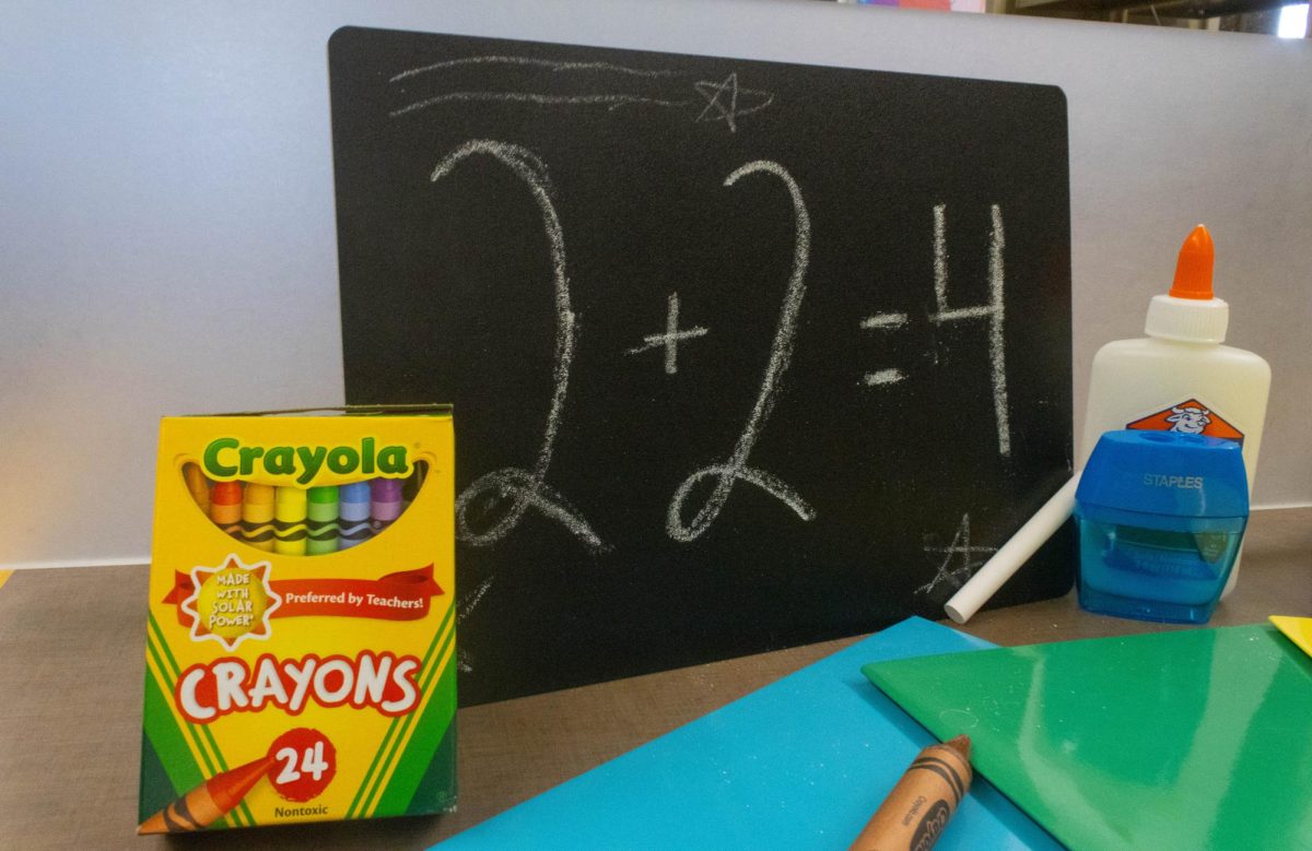 A black chalkboard with "2+2=4" written on it with white chalk stands behind a crayons box and a bottle of glue.