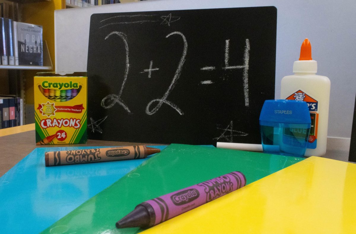 A black chalkboard with the characters "2+2=4" written on it with white chalk stands behind a crayons container, as well as a glue bottle and a pencil sharpener.