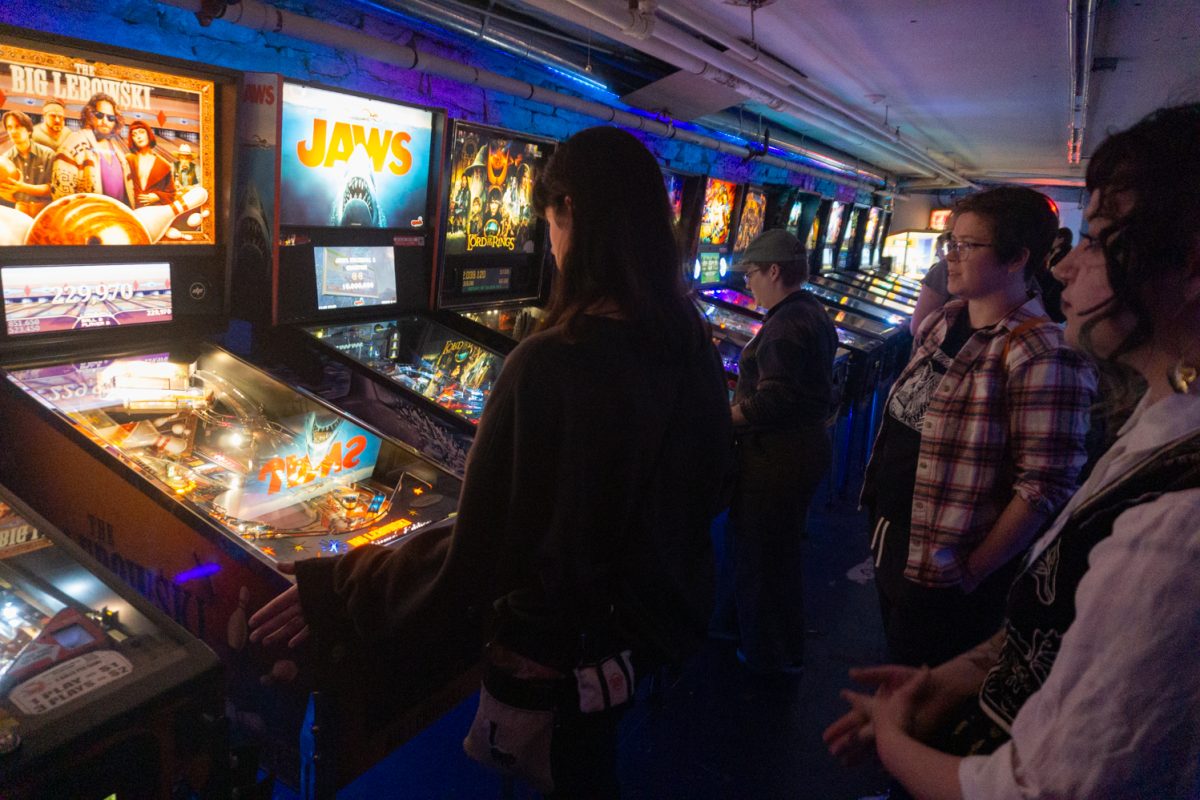 A team watches as a participant plays the "Big Lebowski" machine during the Women, Trans, Femme league at Pinball Jones Feb 12. Ericka Stonebay, who helped establish the league, said the league's goal is to create a space for women, trans, and femme-identifying players in the world of pinball, which is a male-dominated sport.