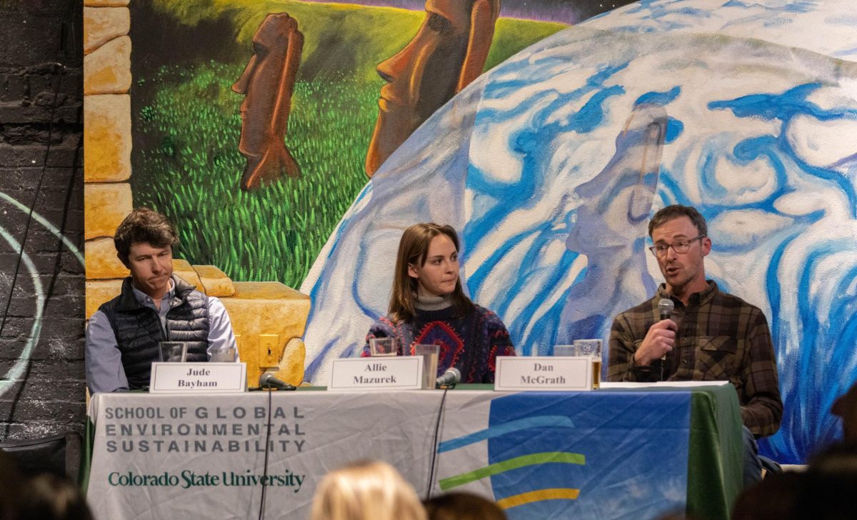 There are two men and one woman sitting at a table, they all have microphones in front of them. A man on the right side of the frame is speaking into the microphone.