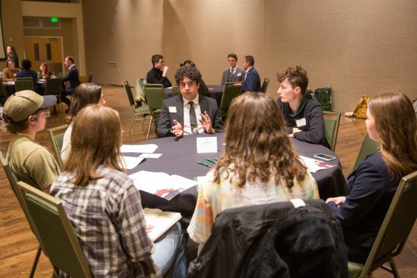 A man in a suit and tie talks at a table amongst people.
