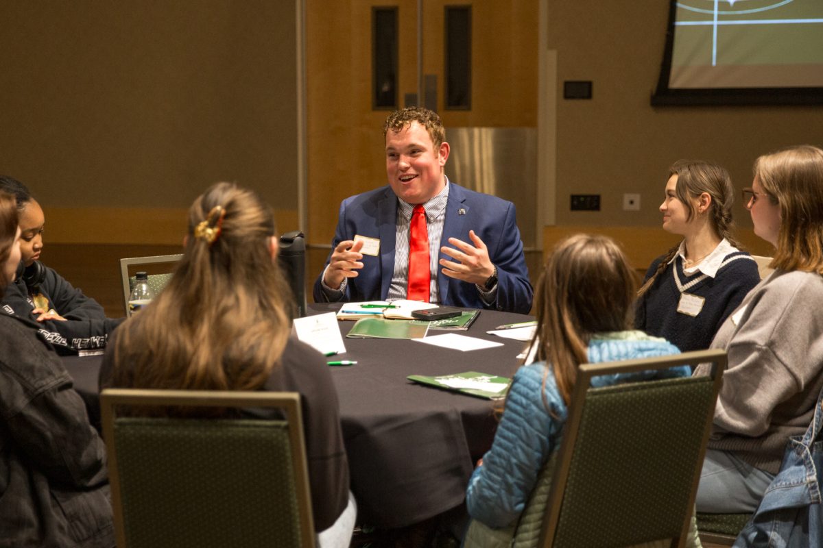 A man wearing a suit and tie sits amongst people talking to them.