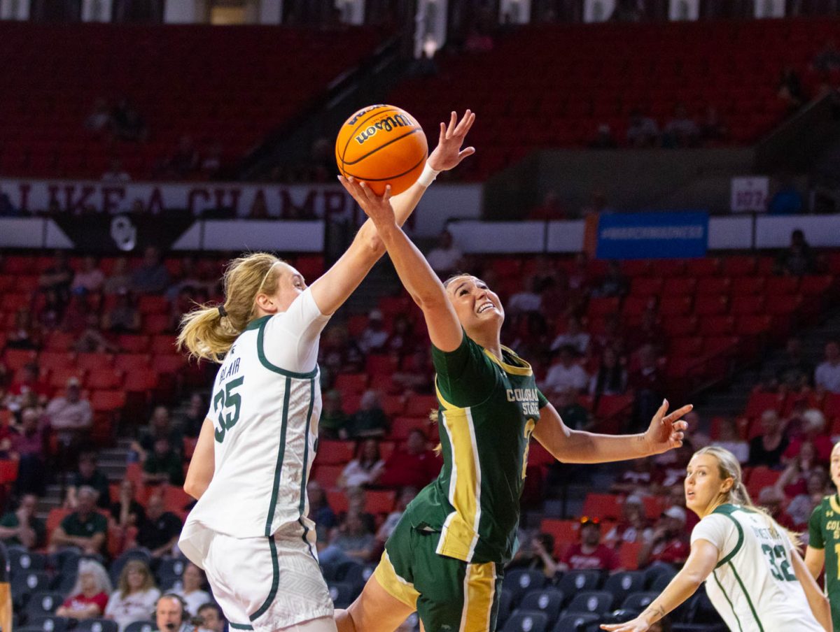 A woman wearing an all green jersey extends her right hand with the ball in it around a woman in an all white jersey.