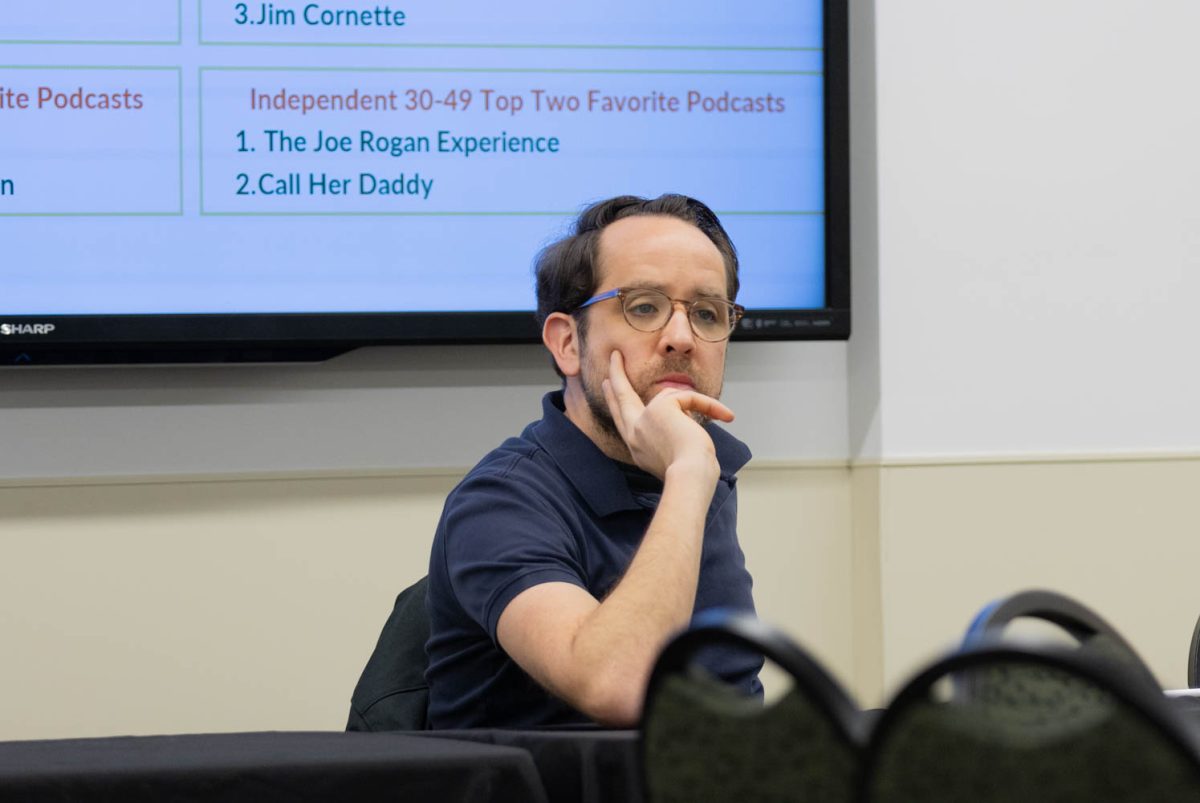 A man in a navy blue polo shirt rests his face in one of his hands attentively.