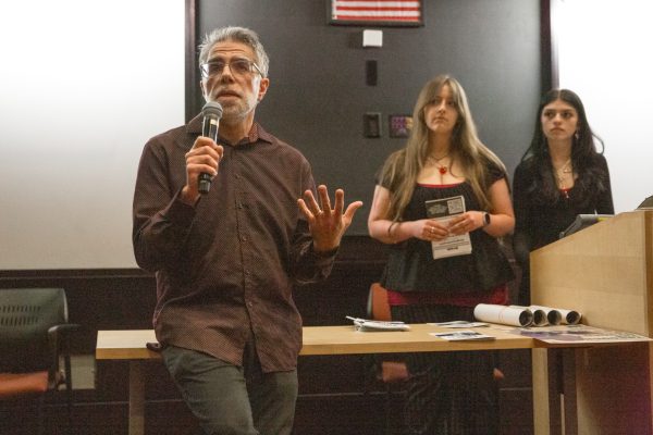 A man in a brown shirt speaks in front of a white and black wall and two women.