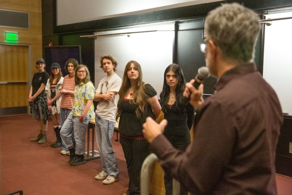 A man with a microphone speaks to eight people looking at him.
