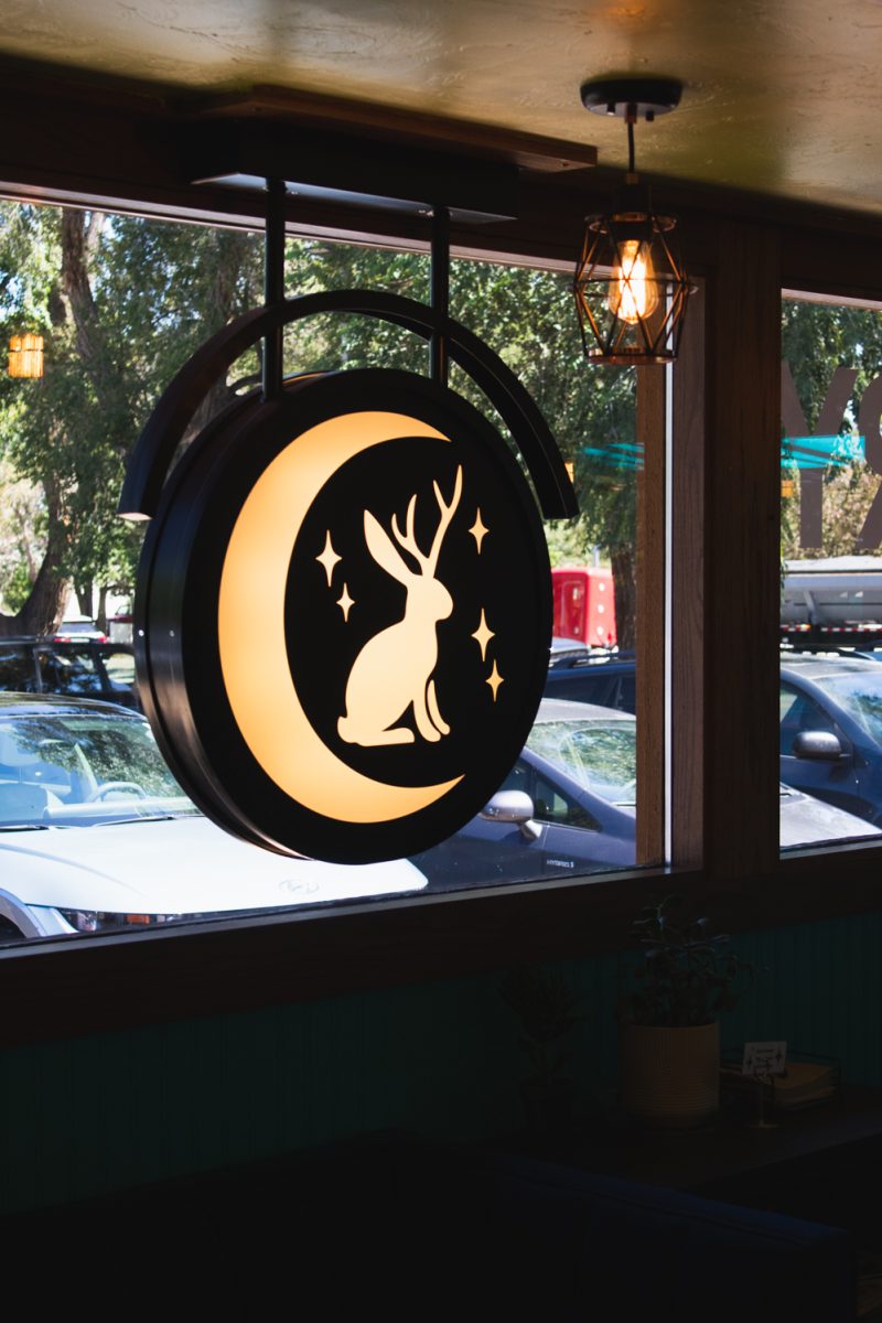 An illuminated sign with the logo of the Trickster Bakery &amp; Cafe hangs in the window Aug. 20. Co-owners Jenn Lewis and Shannon Morr opened the business together, converting it from a pizza hut to a bakery and cafe. “I think both of us are just really kind of proud of the vibe we’re cultivating here,” Morr said, “It’s nice to see all these people coming in and [being] excited about our, like, pride flags on our doors, and all our colorful chairs and couches and stuff, and our big community table.”