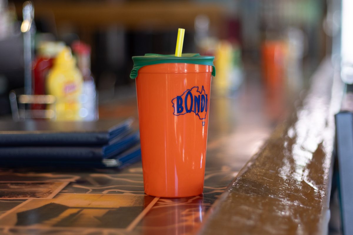A tall orange plastic cup with "BONDI" printed on it sits on a brown counter. The drink cap is green with a yellow straw sticking out of it.